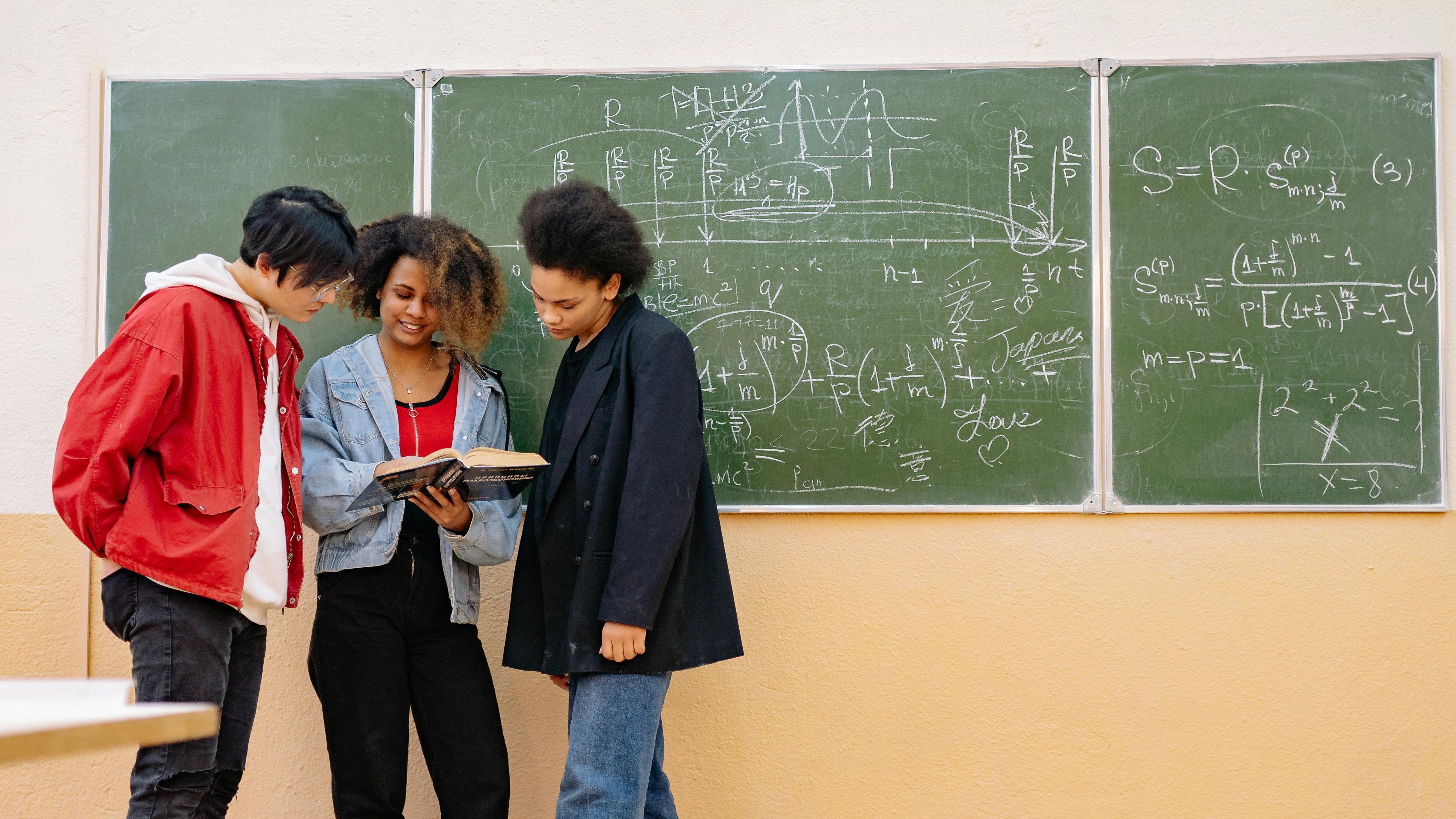 3 Students stand in a corner and look over a textbook with a blackboard in the background